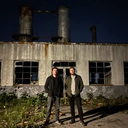 2 guys standing outside of an abandoned broken down derelict jelly factory building with broken windows at night
