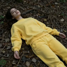 young female, lying dead, covered by dark colored leaves and twigs, in a forest, she is wearing a yellow velor sweat suit