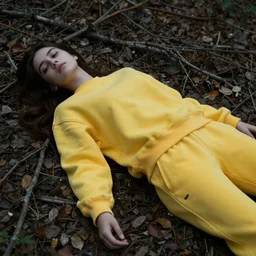 young female wearing a yellow velor sweat suit lying dead on the ground in a forest. She is mostly covered by dark colored leaves and twigs