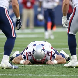patriots qb mac jones laying face down on the field as the opposing team celebrates a touchdown in the endzone in the background