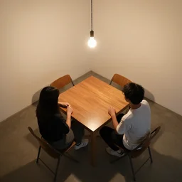 overhead view of two people sitting across from each other at a table in a bare room with a single hanging light bulb above them, the table is square wood table and they are sitting on folding chairs