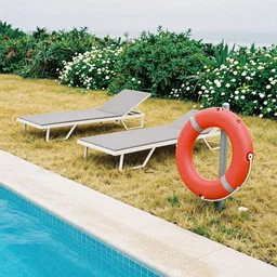 two tellow beach beds and a red plastic life saver next to a pool, yellowish grass and green bushes enviorment with white flowers, grainy photograph, diagonal composition, cloudy day, long shot, close up