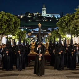 A Holy Week procession of souls in a lush Spanish landscape at night. A figure at the front holds a cross. Art Deco.