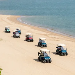 wide shot of a parade of 1 0 golf carts driving down shoreline of sandy beach next to the water. no grass. no trees
