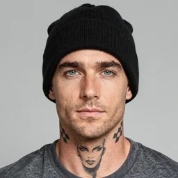 man in black beanie hat. Man has blue eyes and a neutral expression. Man appears to be in 30s or 40s. Man has bushy, groomed eyebrows. On man's neck side is a tattoo of a checkered flag. On man's neck center is a tattoo of a female face. Man is wearing gray t-shirt. Man has light stubble. Photograph from shoulders-up.
