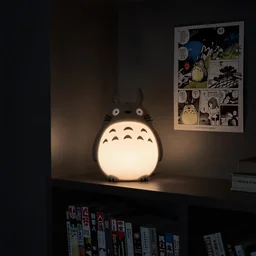 a lamp that is placed in a shelf in a dark room. the lamp is shaped like totoro. the room is dark. the lamp lights the room. the shelf holds comic books