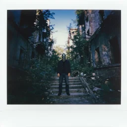 A polaroid photo of a mysterious man with a gas mask standing in the midle of a staircase alley looking in the direction of the camera :: exterior, blue sky visible :: ruined city with vegetation and trees growing everywhere on the destroyed buildings :: forest :: apocalyptic, gloomy, desolate :: long shot, low angle, dramatic backlight, symmetrical, night, slightly colorful photography :: cinematic shot, very detailed