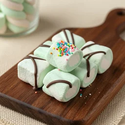 photograph of a marshmallow cube with sprinkles on a dark wooden chopping board, chocolate drizzle, green pastel colours, hessian cloth, styled food photography, photorealistic, 4 k