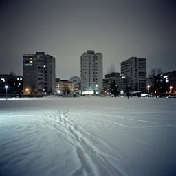 a snow covered field with buildings in the background, a matte painting by zlatyu boyadzhiev, featured on unsplash, brutalism, nightscape, quantum wavetracing, photo taken with ektachrome