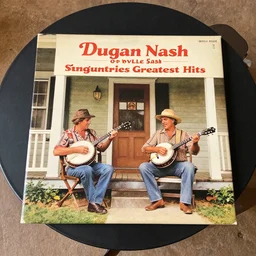 a dust vinyl record album cover of dugan nash and orville sash sing countries greatest hits with two hillbillies playing banjos on a porch : the album cover is on a coffee table
