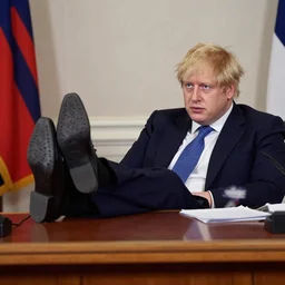 Boris Johnson wearing suit and necktie and black shoes resting his feet up on a desk