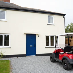 a detached house in england with offwhite render and a blue alumium front door. Grey gravel drive and a red ve golf car in the drive