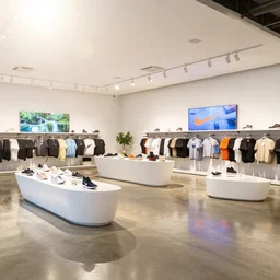 1 8 mm photograph atmospheric, realistic lighting, light bloom sunlight, nature outside, reflections, large nike store. white walls. polished concrete floor. display tables with shoes and clothes. low ceilings with downlights and spots. curved white furniture with large digital screens. a few plants in the background