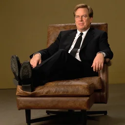 2 0 0 3 john lasseter wearing a black suit and necktie and black shoes sitting in a chair with his legs resting on top of a footstool.
