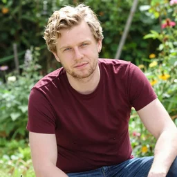 photographic portrait of a polish man, 37 years old, short blonde hair in loose curls, small blue eyes, very pale white skin, medium blond stubble, wearing a maroon tight t-shirt and blue jeans, an earring on left ear, thin lips, smirking, posing in a garden, under bright sunlight