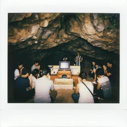 A polaroid photo of a group of people inside a wide cave praying to a wooden computer on a altar