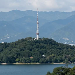 a hill with a radio tower next to a lake, mountain range in background. telephoto lens photography.