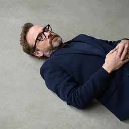 bryan cranston with a beard, black reading glasses while laying on a smooth concrete floor